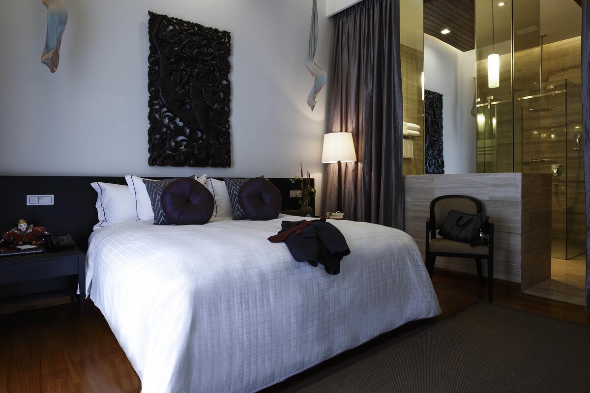 A hotel room with a large bed, a nightstand, a lamp, a chair, and a glass shower area.