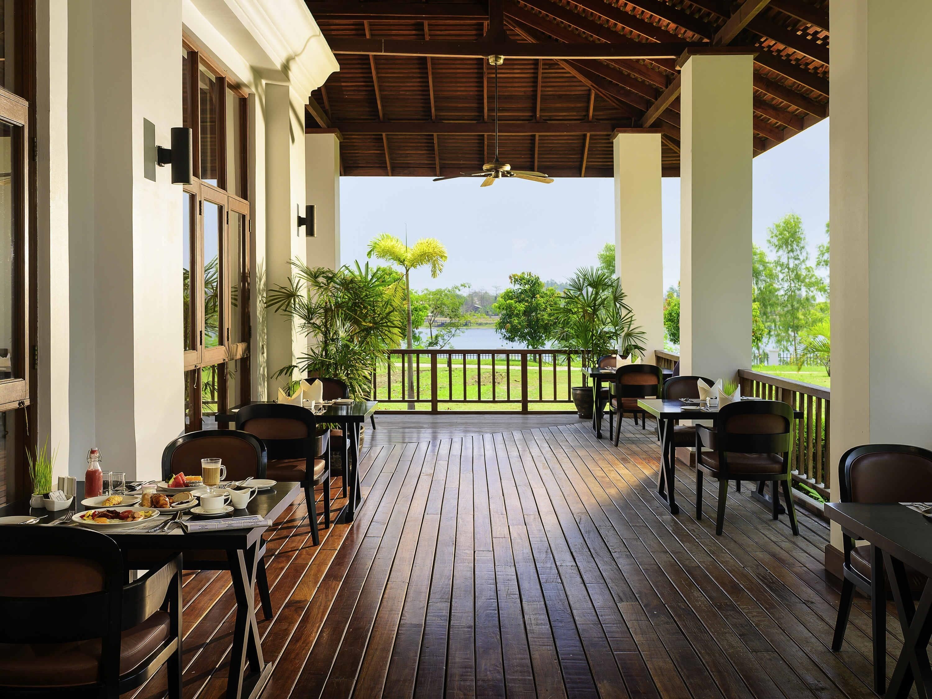 Un espacio de comedor al aire libre cubierto, con suelo de madera, mesas, sillas y vistas a un lago.