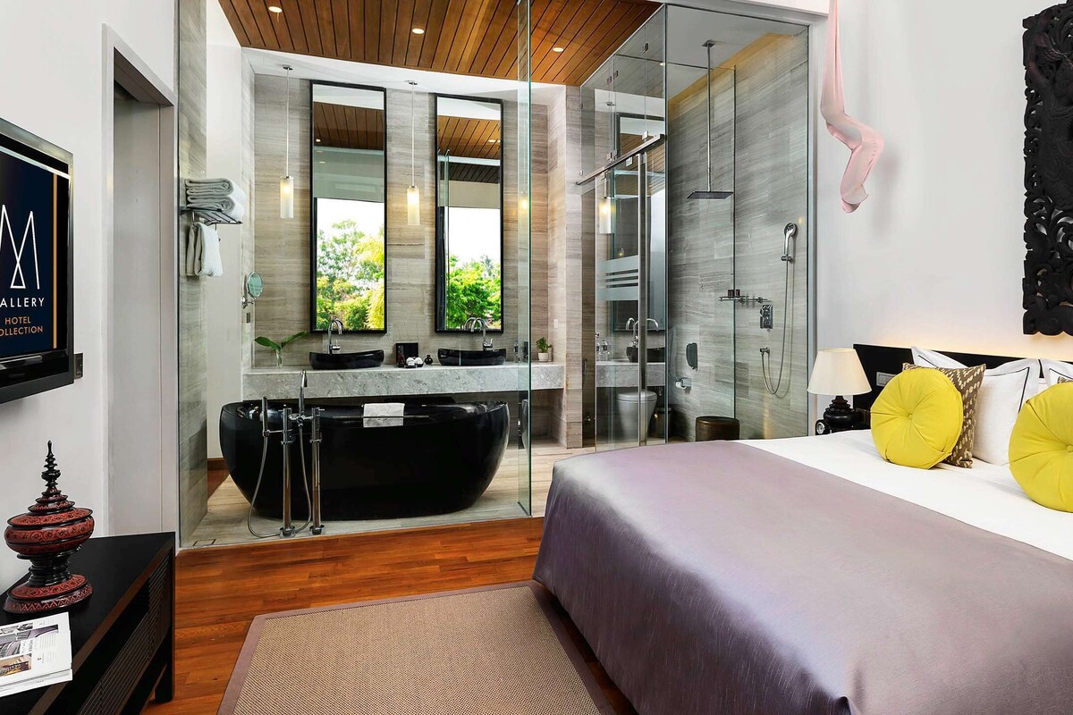 Une chambre d’hôtel moderne avec un grand lit, une baignoire noire, une douche séparée par des parois de verre et une vue sur la verdure par les fenêtres.