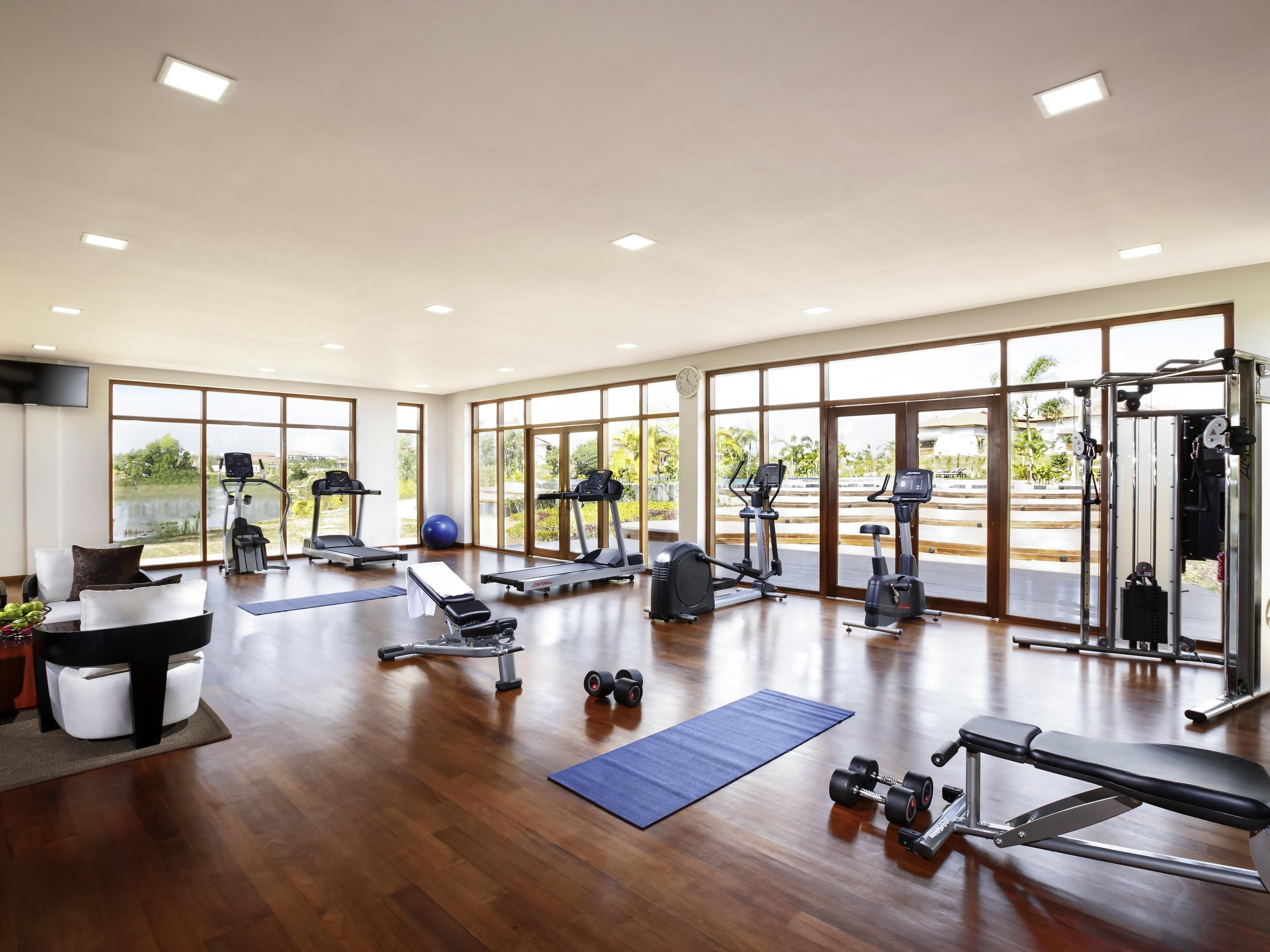 A spacious gym with various exercise equipment, large windows, and a wooden floor.