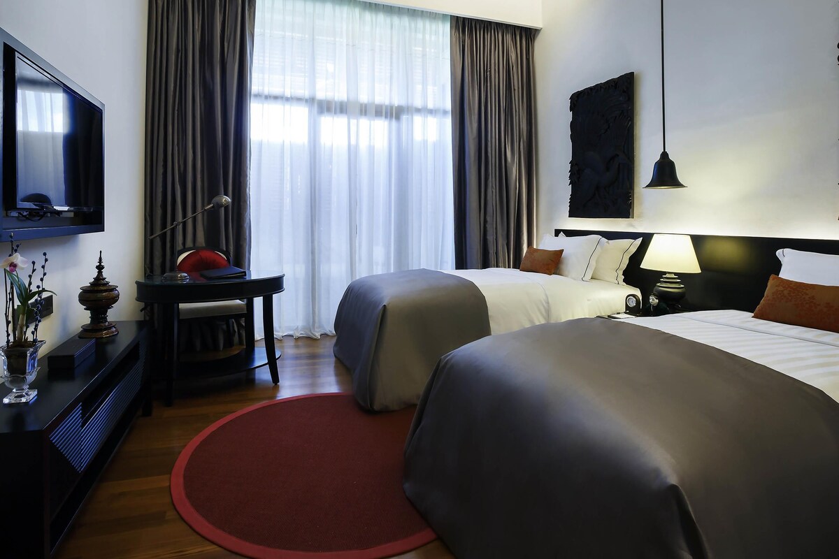 A hotel room with two beds, a desk with a lamp, a TV, and a large window with curtains.