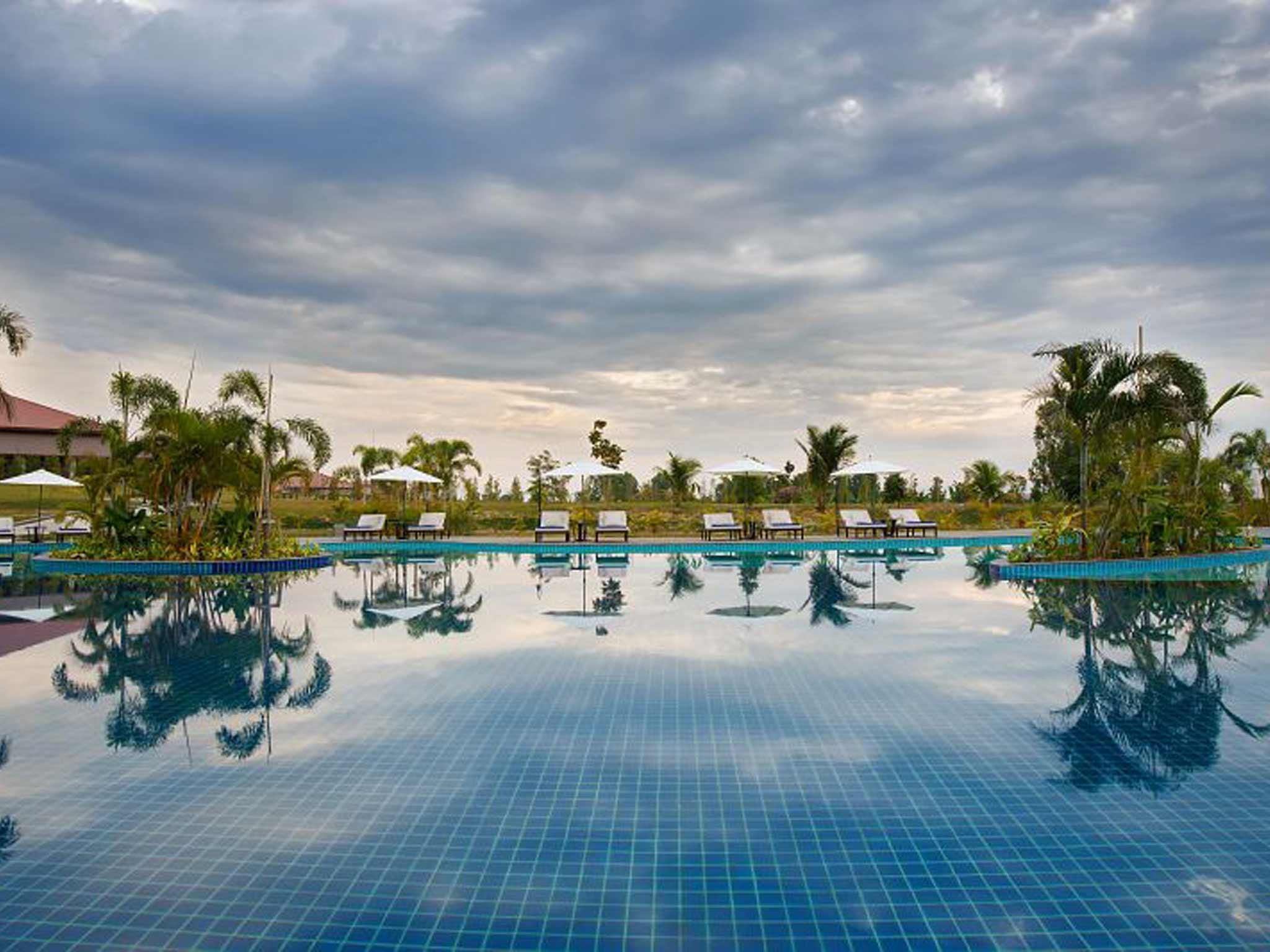 Una piscina amplia y cristalina con sillas de descanso y sombrillas, rodeada de palmeras y un cielo nublado.