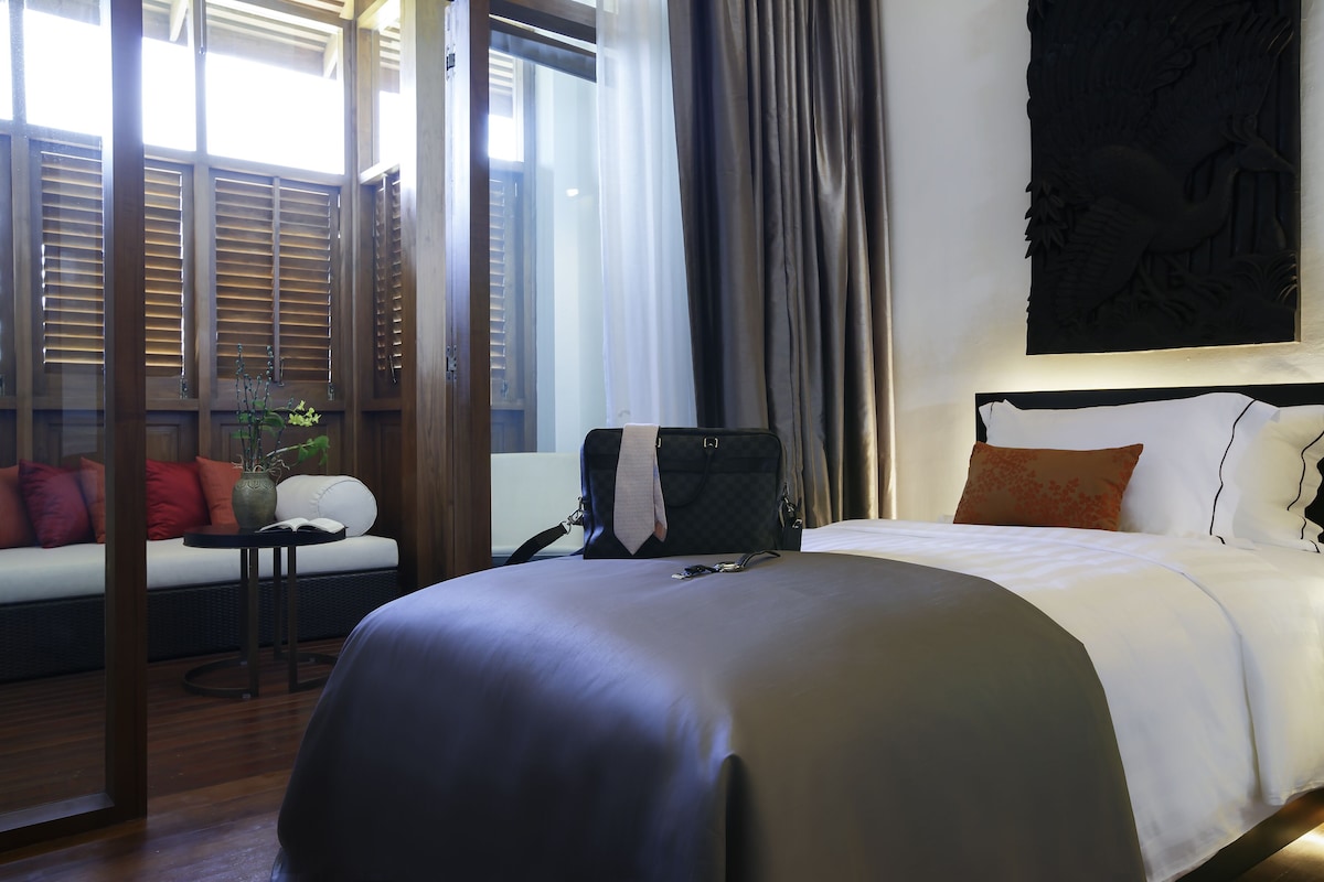 Une chambre d’hôtel avec un lit, une petite table sur laquelle est posé un vase, et un canapé avec des coussins rouges.