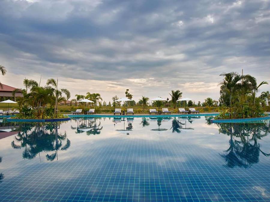 Una piscina amplia y cristalina con sillas de descanso y sombrillas, rodeada de palmeras y un cielo nublado.