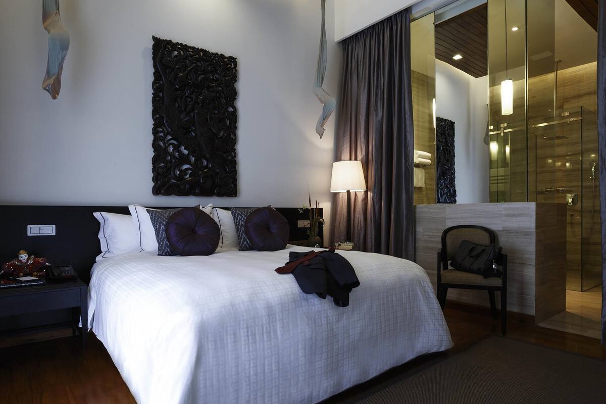 Una habitación de hotel con una cama grande, una mesita de noche, una lámpara, una silla y un área de ducha con mampara de vidrio.