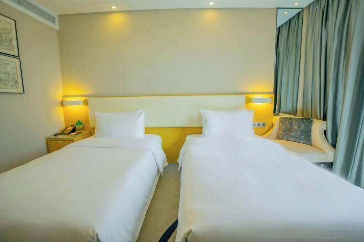 Una habitación de hotel con dos camas individuales, una mesita de noche con lámpara y un gran ventanal con cortinas.