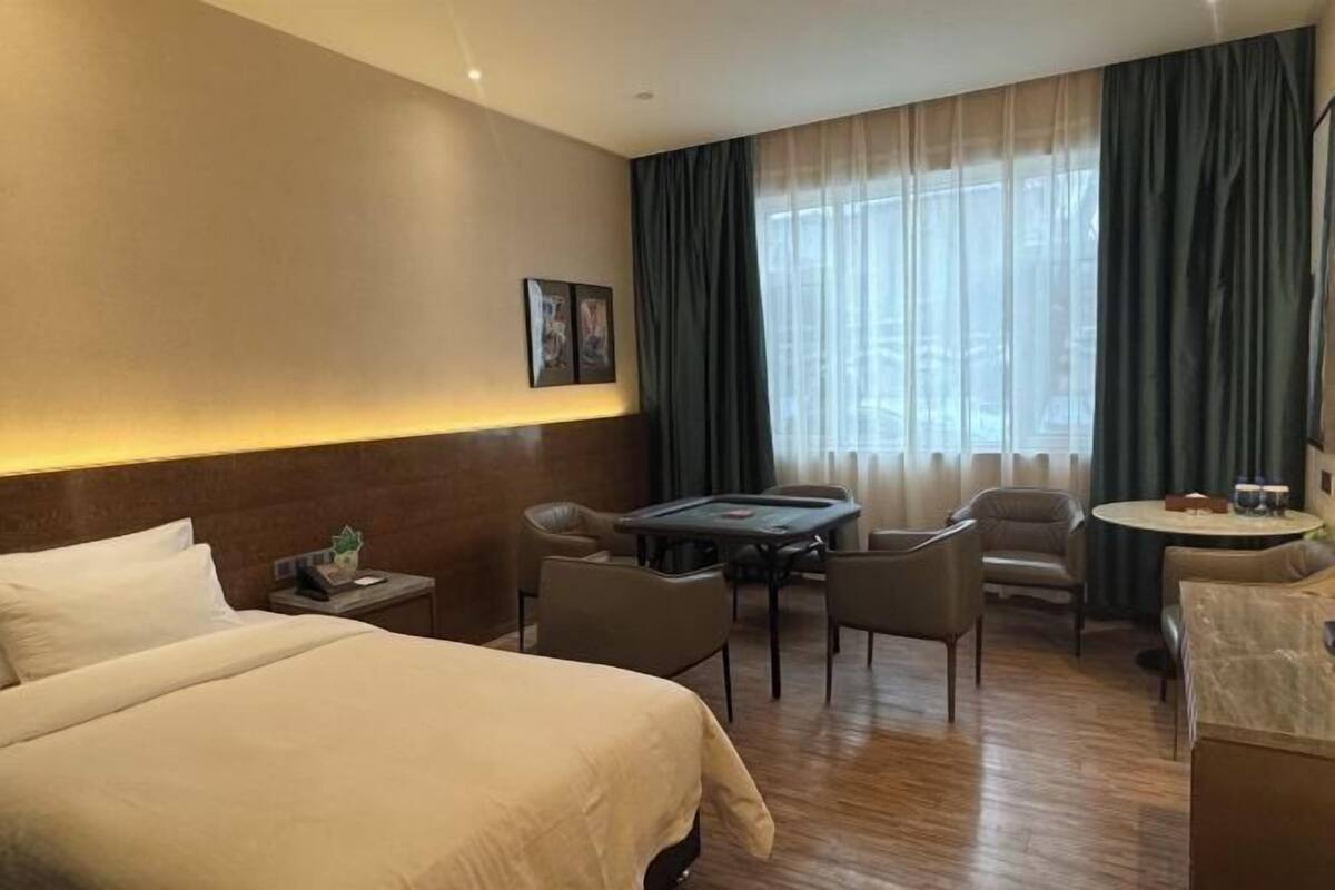 Habitación de hotel con una cama grande, un escritorio con sillas, una mesita y vistas al exterior a través de cortinas translúcidas.