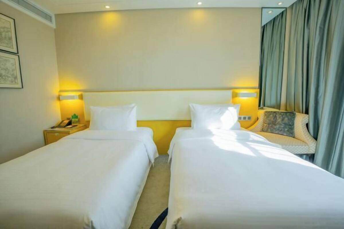 Habitación de hotel con dos camas individuales, una mesita de noche con lámpara y un ventanal con cortinas.