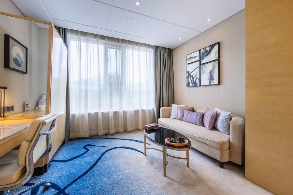 Ein Hotelzimmer mit einer Couch, einem Couchtisch, einem Schreibtisch mit Stuhl und einem großen Fenster mit Vorhängen.