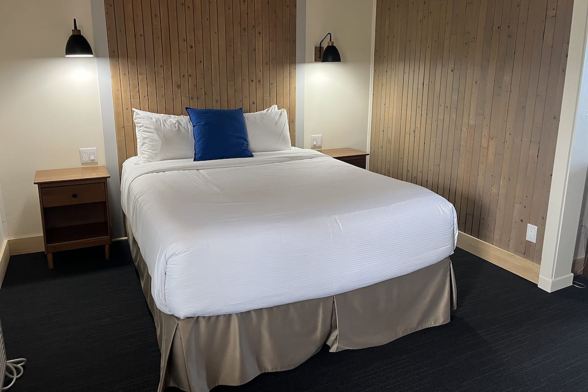 A neatly made bed with a white duvet and a blue pillow, flanked by two bedside tables with lamps, in a room with wooden paneling on the walls and a dark carpeted floor.