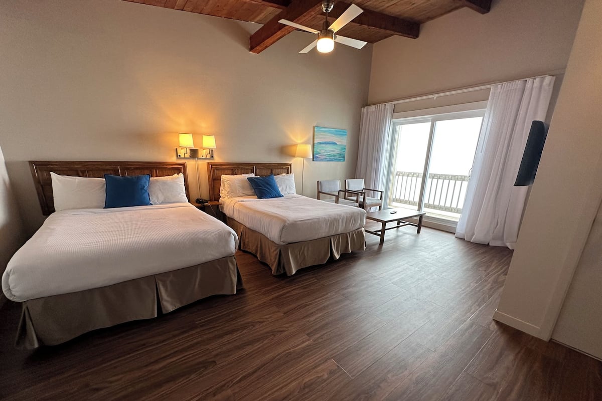A hotel room with two beds, a small table, chairs, a ceiling fan, and a large window with curtains.