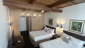 A hotel room with two beds, wooden ceiling beams, a door, and a wall-mounted light fixture. - Hôtel Motel La Côte Surprise (Perce)