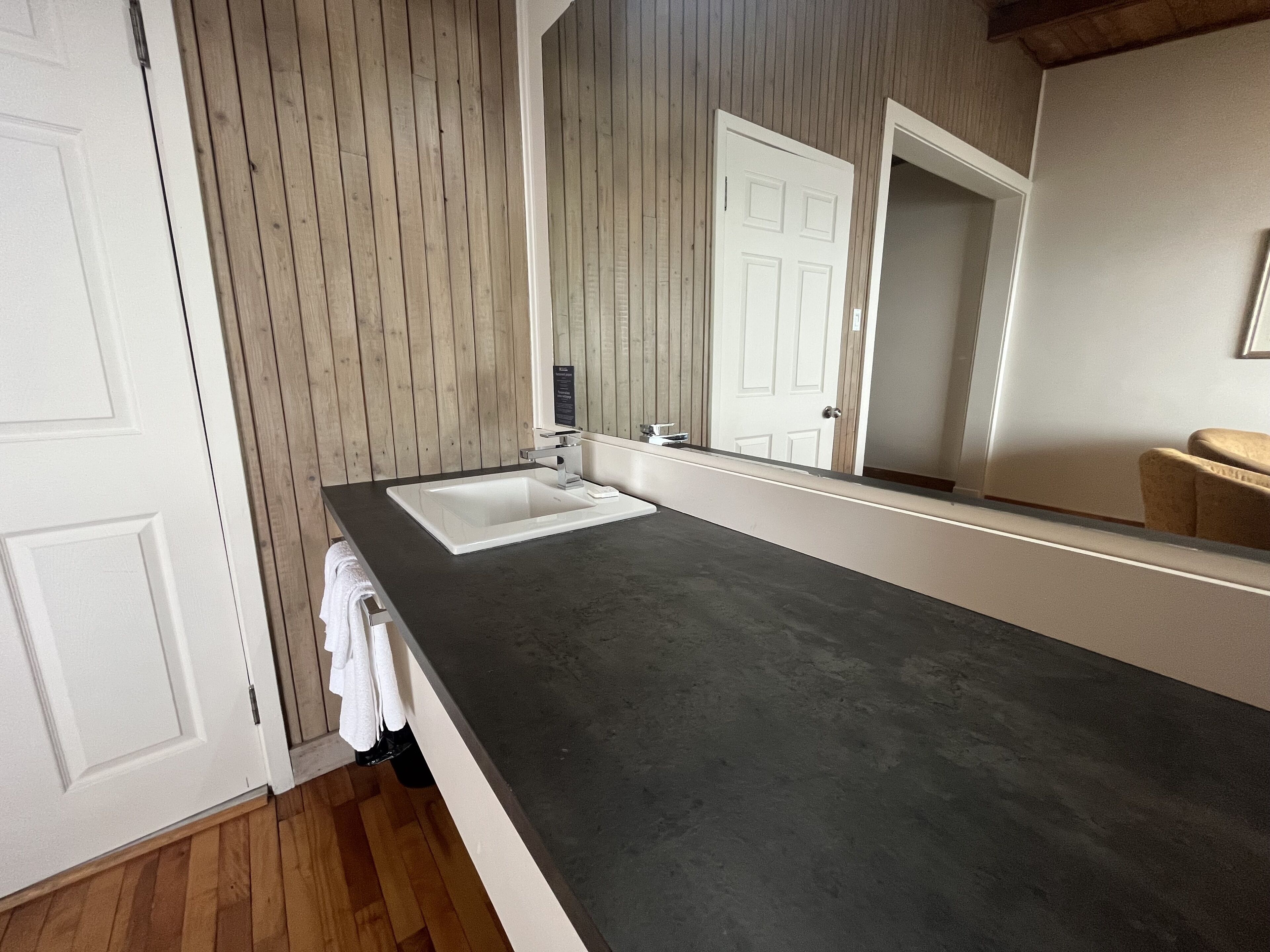 A bathroom with a white sink, wooden panel walls, a large mirror, and a dark countertop.