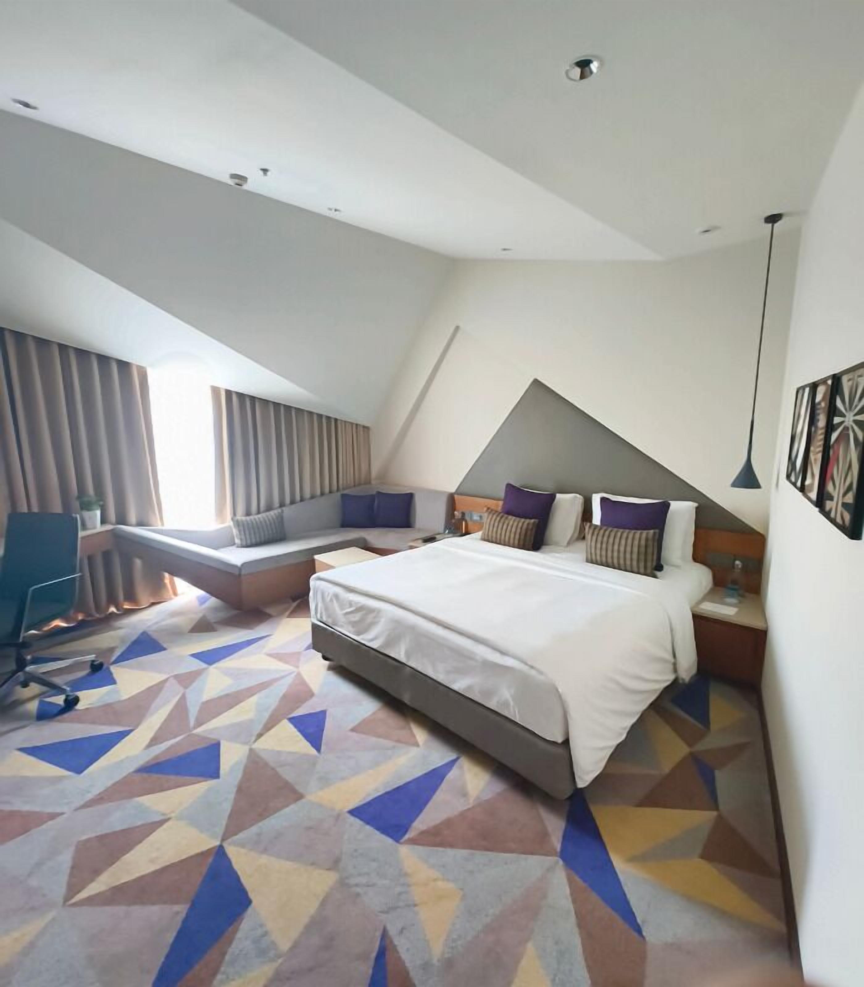 A modern hotel room with a large bed, a seating area, and a geometric patterned floor.