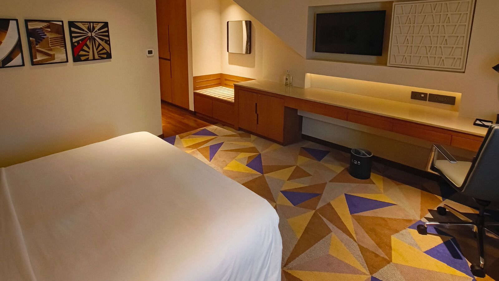 A hotel room with a bed, a desk with a chair, a TV, and a patterned carpet.