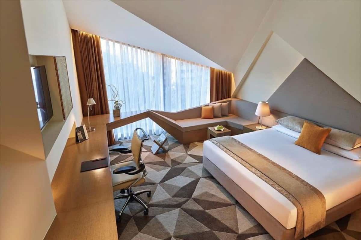 A modern hotel room with a bed, desk, chair, and a view of the city through large windows.