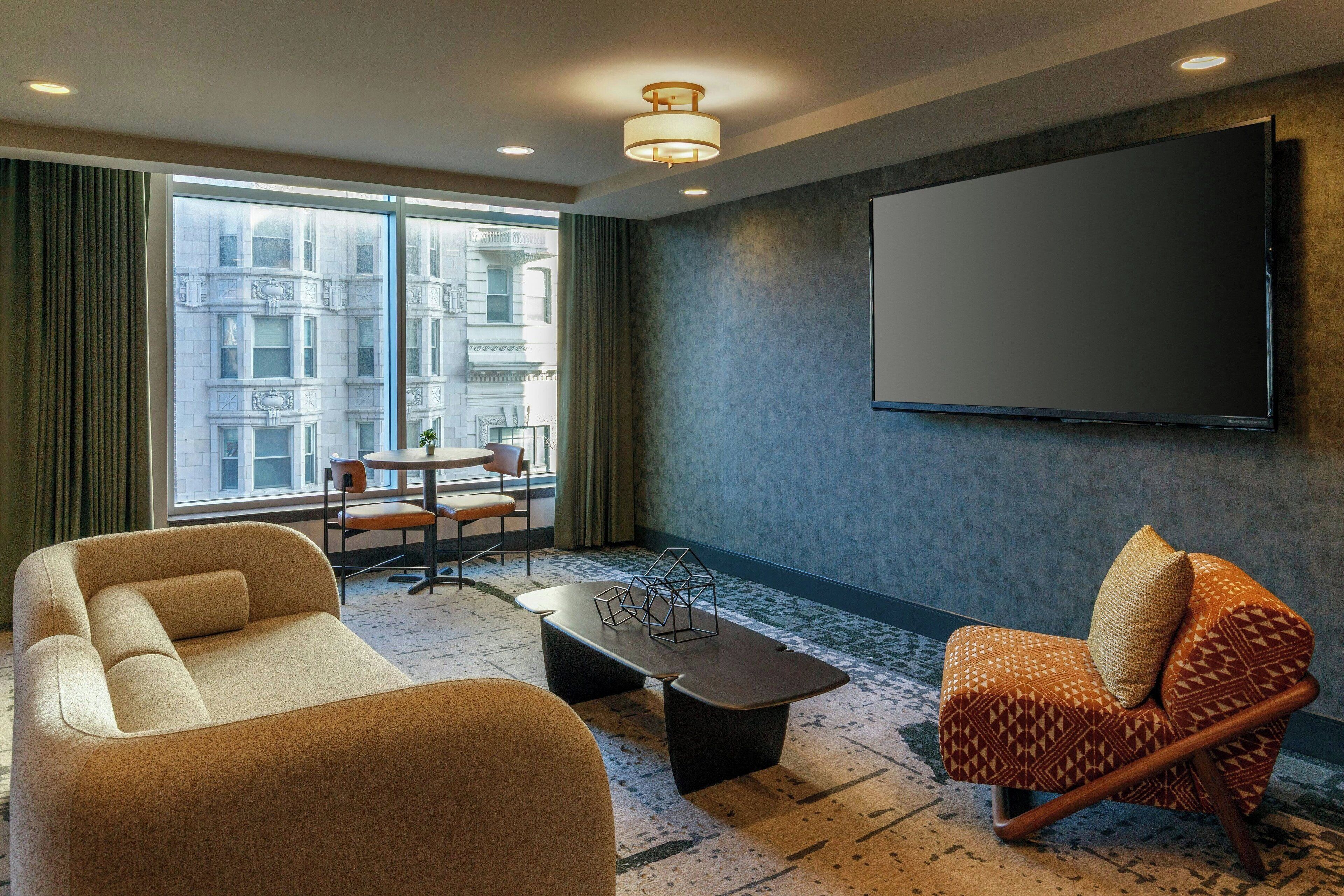 Una sala de estar moderna con sofá, sillón y mesa de centro. Un televisor de pantalla plana grande está montado en la pared, y hay una ventana con vistas a edificios.
