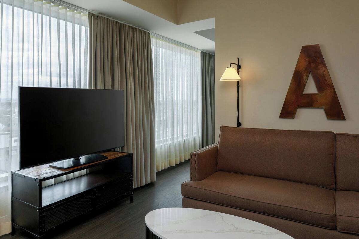 A modern living room with a flat-screen TV on a wooden entertainment unit, a brown leather sofa, a white marble coffee table, and a large letter 'A' on the wall.