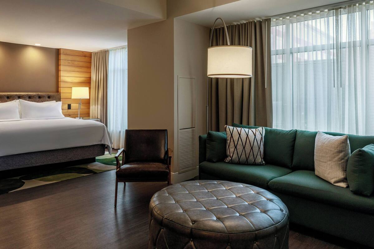A hotel room with a bed, a green sofa, a leather ottoman, a chair, and a lamp.