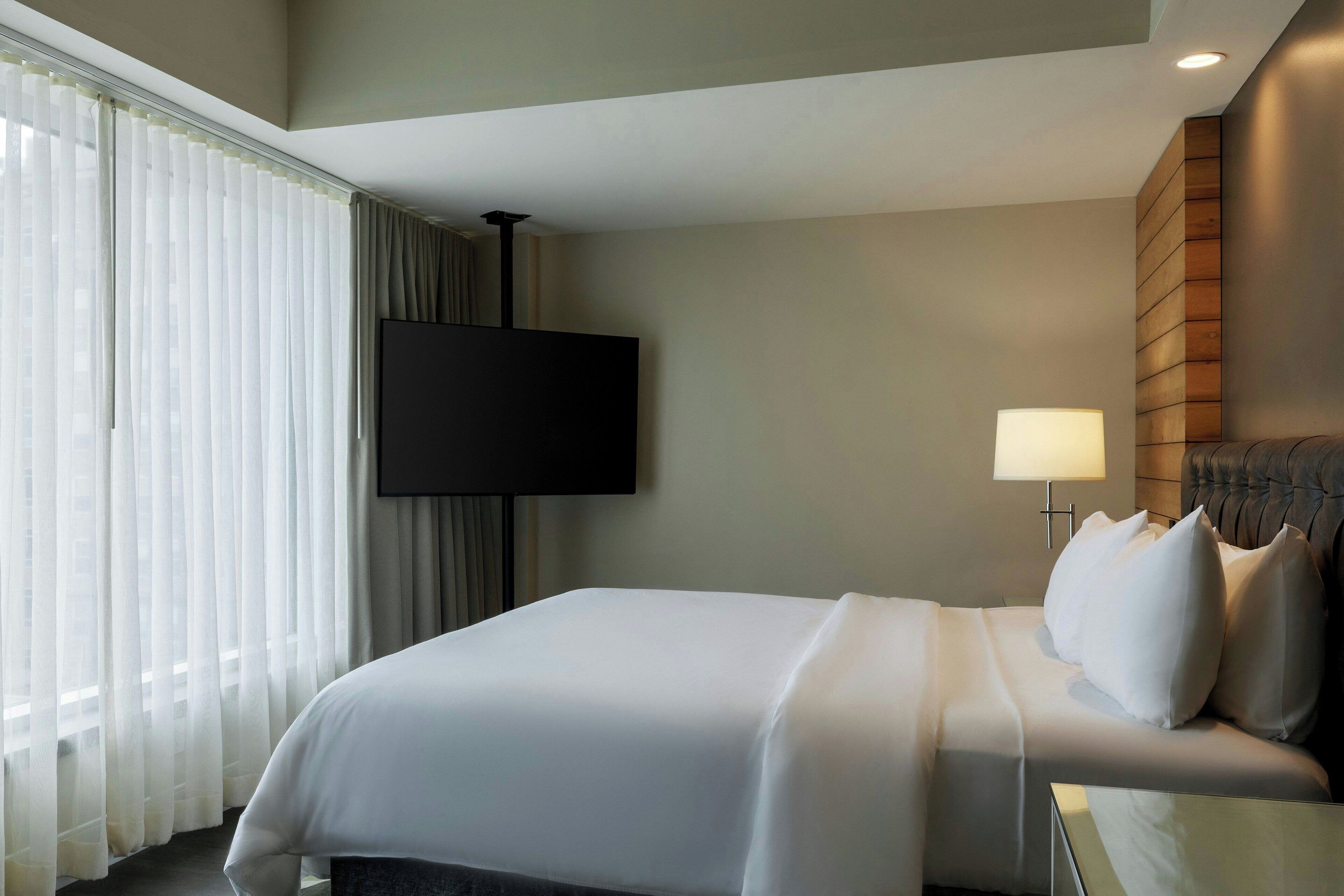 A hotel room with a bed, a TV mounted on the wall, a bedside lamp, and a window with sheer curtains.
