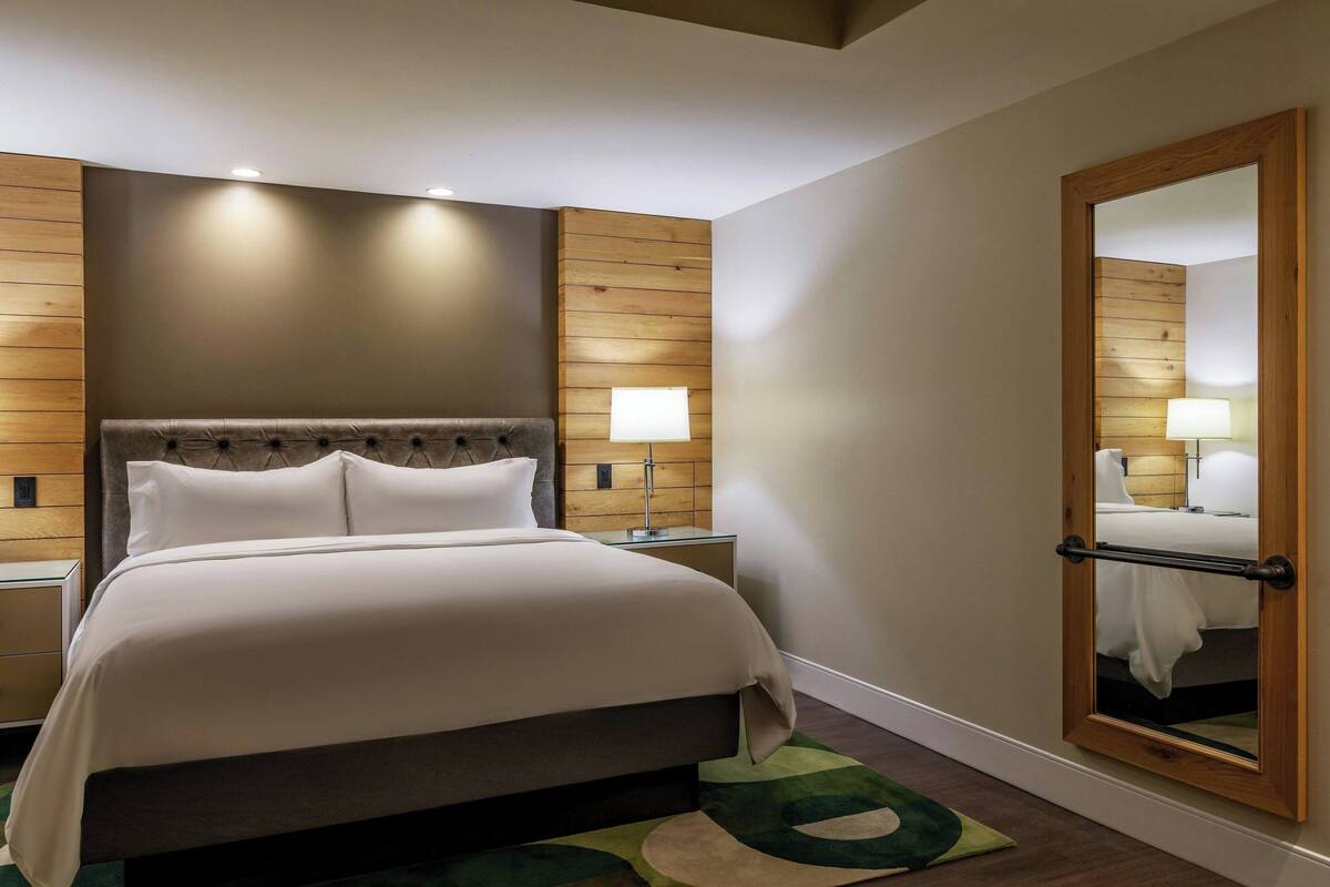 A hotel room with a large bed, wooden panel walls, a mirror, and a nightstand with a lamp.