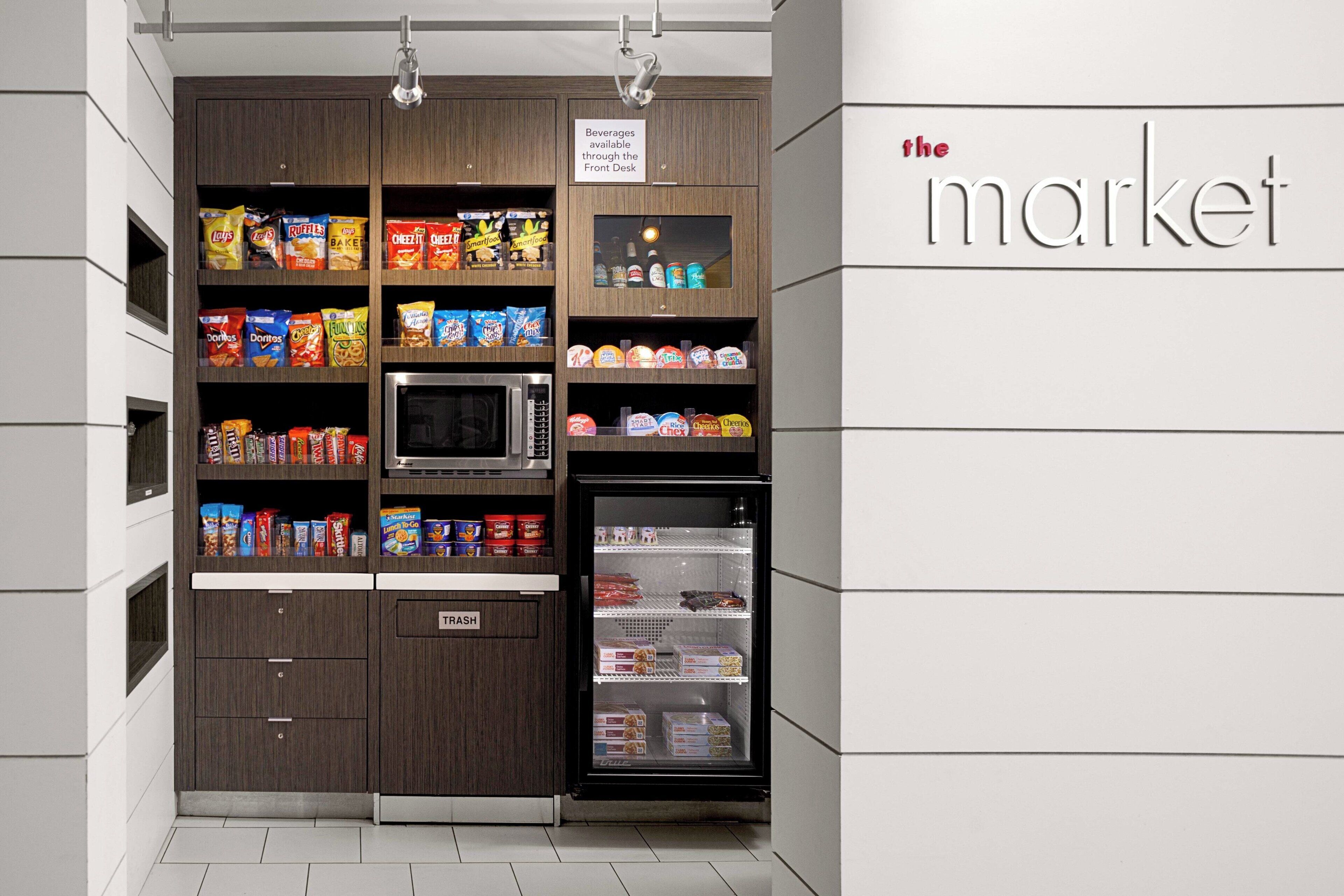 Una zona de cocina con microondas, refrigerador y estantes llenos de aperitivos y bebidas. Un letrero indica que las bebidas están disponibles en la recepción.