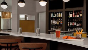 Zona de bar con mostrador blanco, taburetes, una bebida y un plato de comida.