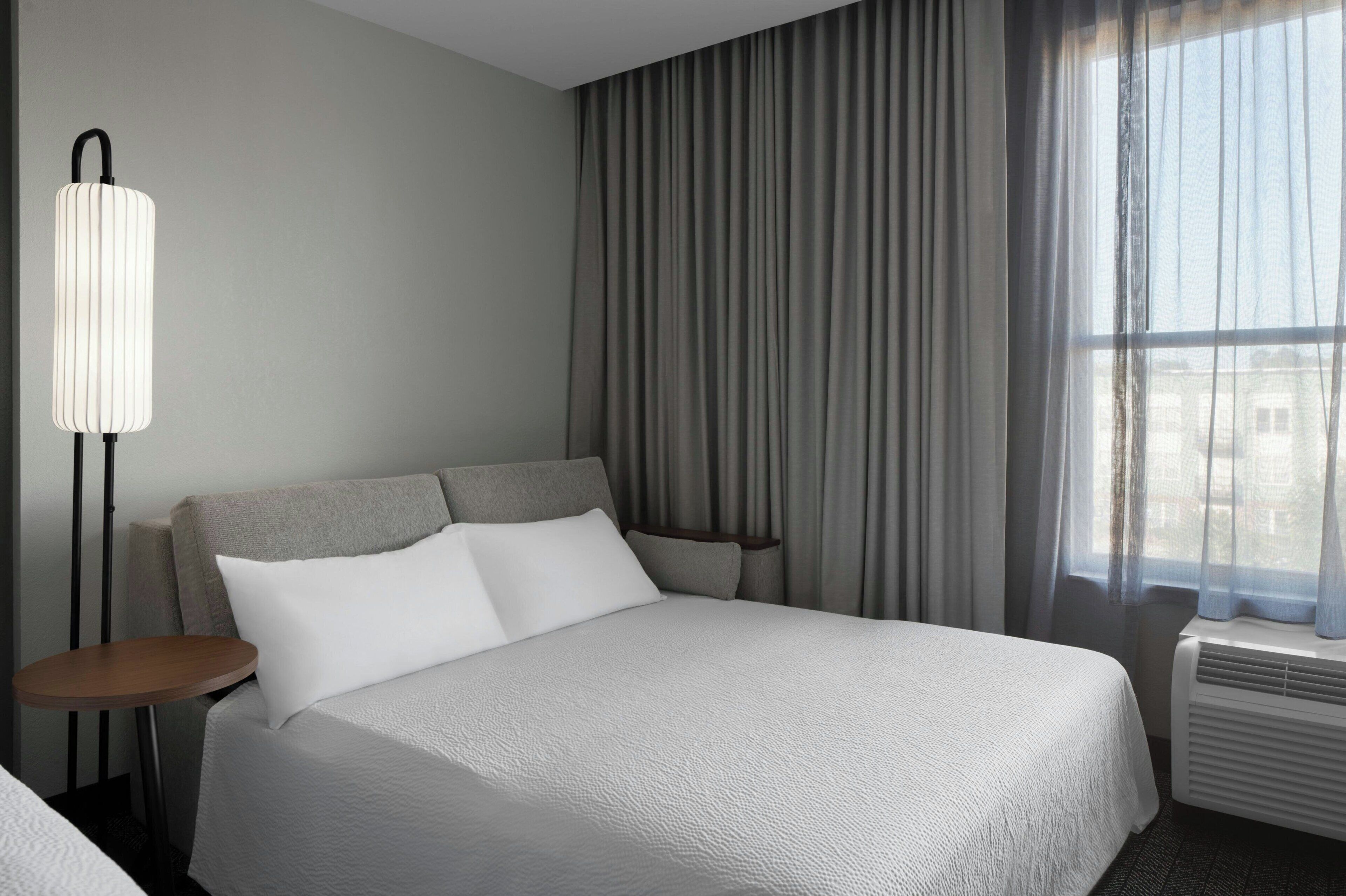 A hotel room with a large bed, a bedside table, a lamp, and a window with curtains.