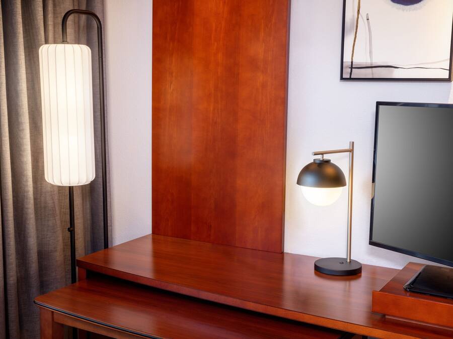 Een houten bureau met een flatscreen televisie, een zwarte bureaulamp en een witte lamp met een geribbelde kap.