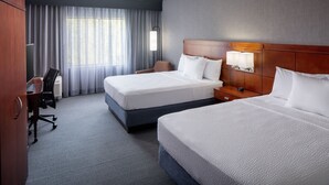 A hotel room with two beds, a desk, a chair, a lamp, and a view of the outdoors through large windows.