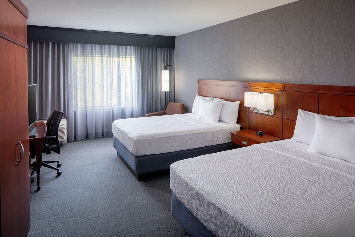 Une chambre d’hôtel avec deux lits, un bureau, une chaise, une lampe et une vue sur l’extérieur grâce à de grandes fenêtres.
