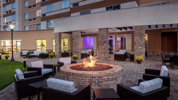 A courtyard with a circular fire pit surrounded by wicker furniture, a brick wall, and a building with multiple windows and a flat roof.