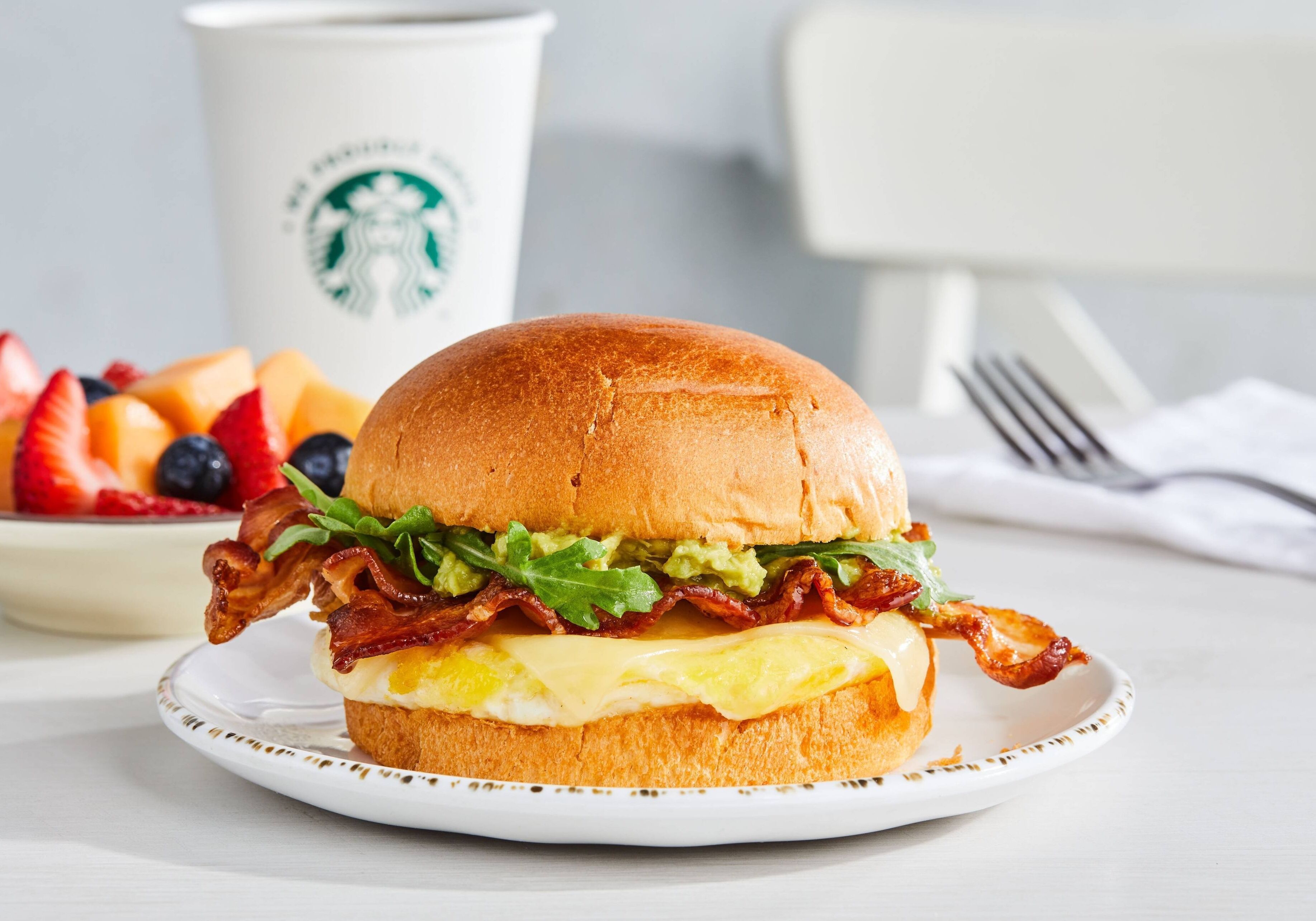 Un sándwich de desayuno en un plato, con una taza y un tenedor al fondo.