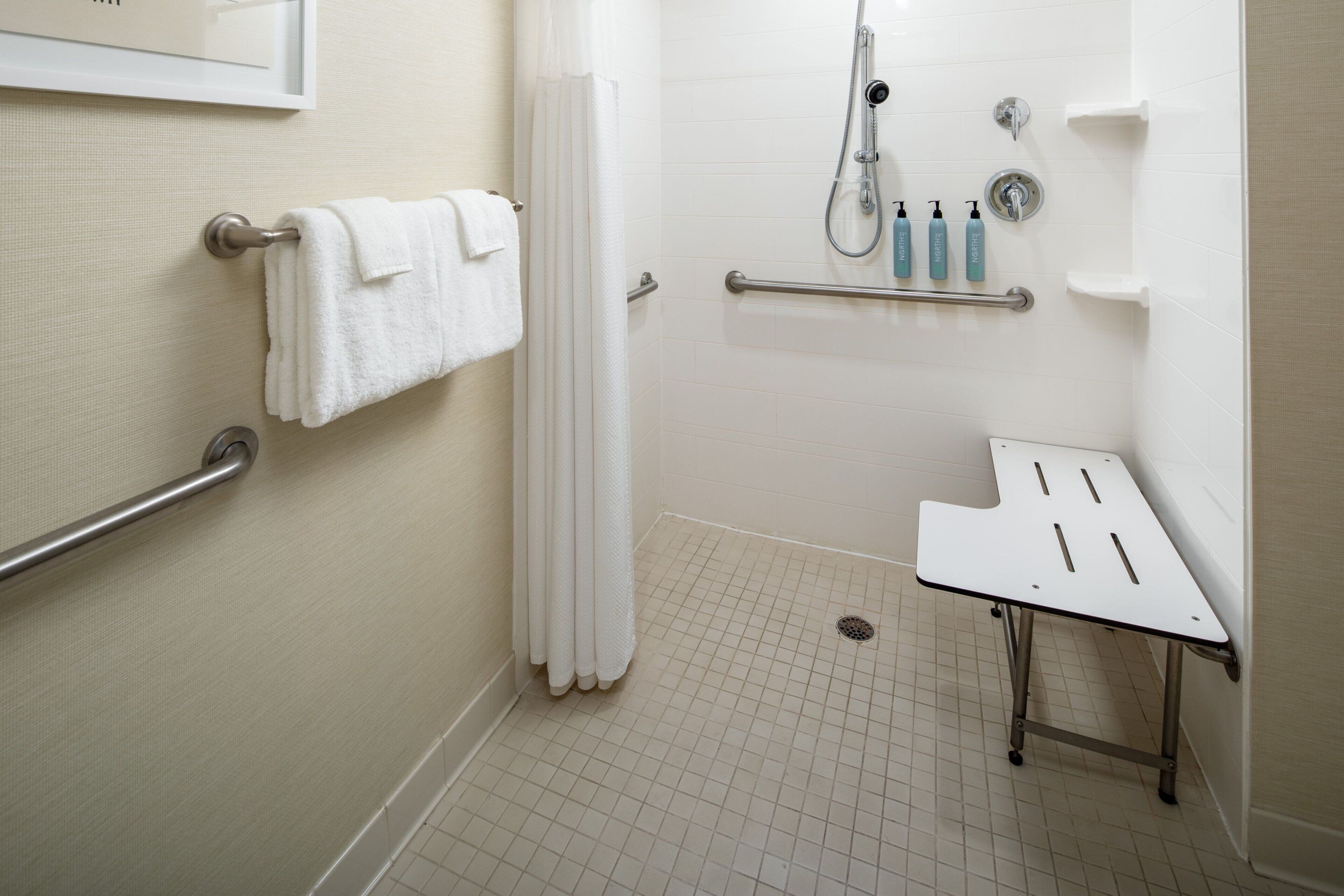 A modern bathroom with a shower, grab bars, a towel rack, and a small table.