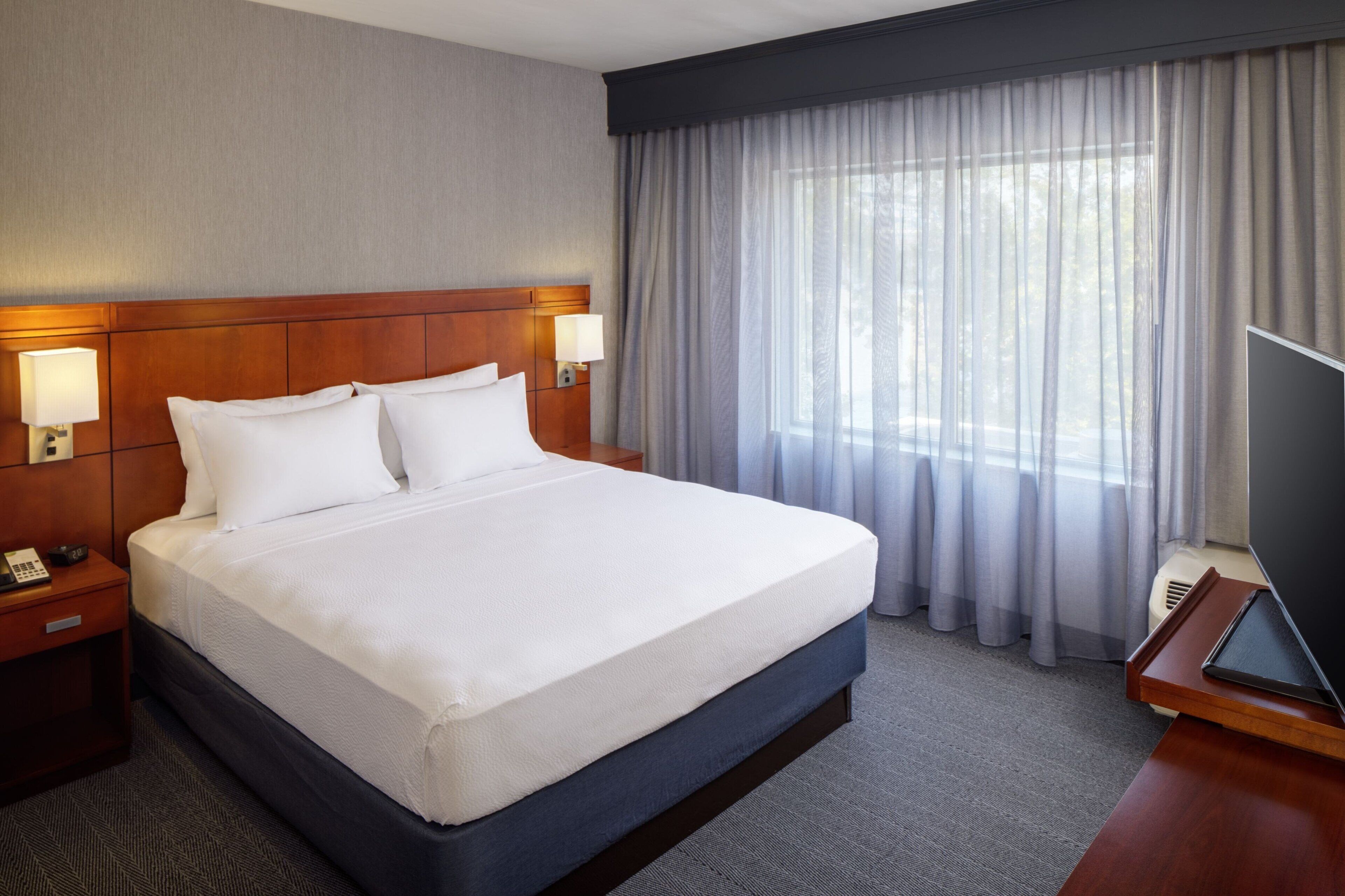 A hotel room with a large bed, wooden headboard, two bedside lamps, a flat-screen TV on a wooden stand, and a window with sheer curtains.