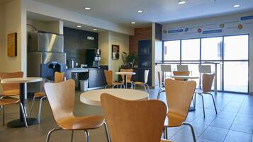 A modern cafe with round tables and wooden chairs, a coffee machine, and a large window offering a view.