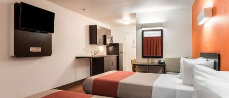 A hotel room with a bed, a desk, a chair, a TV mounted on the wall, and a kitchenette.