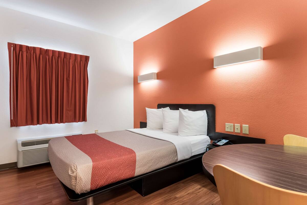 Une chambre d’hôtel avec un lit, une table et une chaise. Il y a une fenêtre avec des rideaux, un luminaire fixé au mur et un radiateur.