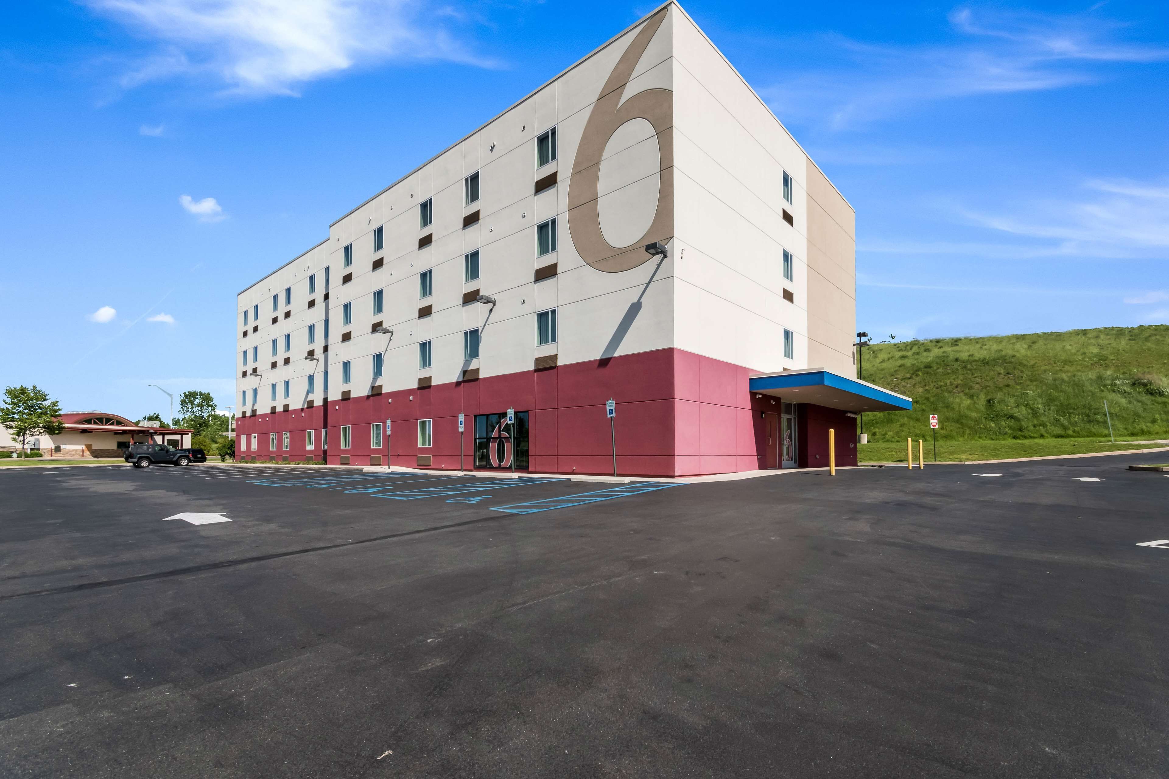 A modern hotel with a large '6' on the facade, a parking lot, and a green hill in the background.