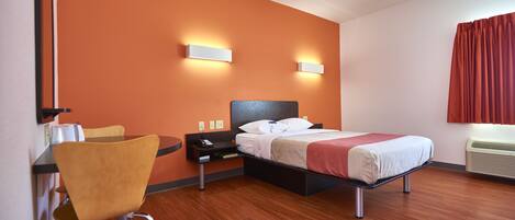 A hotel room with a bed, a chair, a desk, and a wall-mounted air conditioner.
