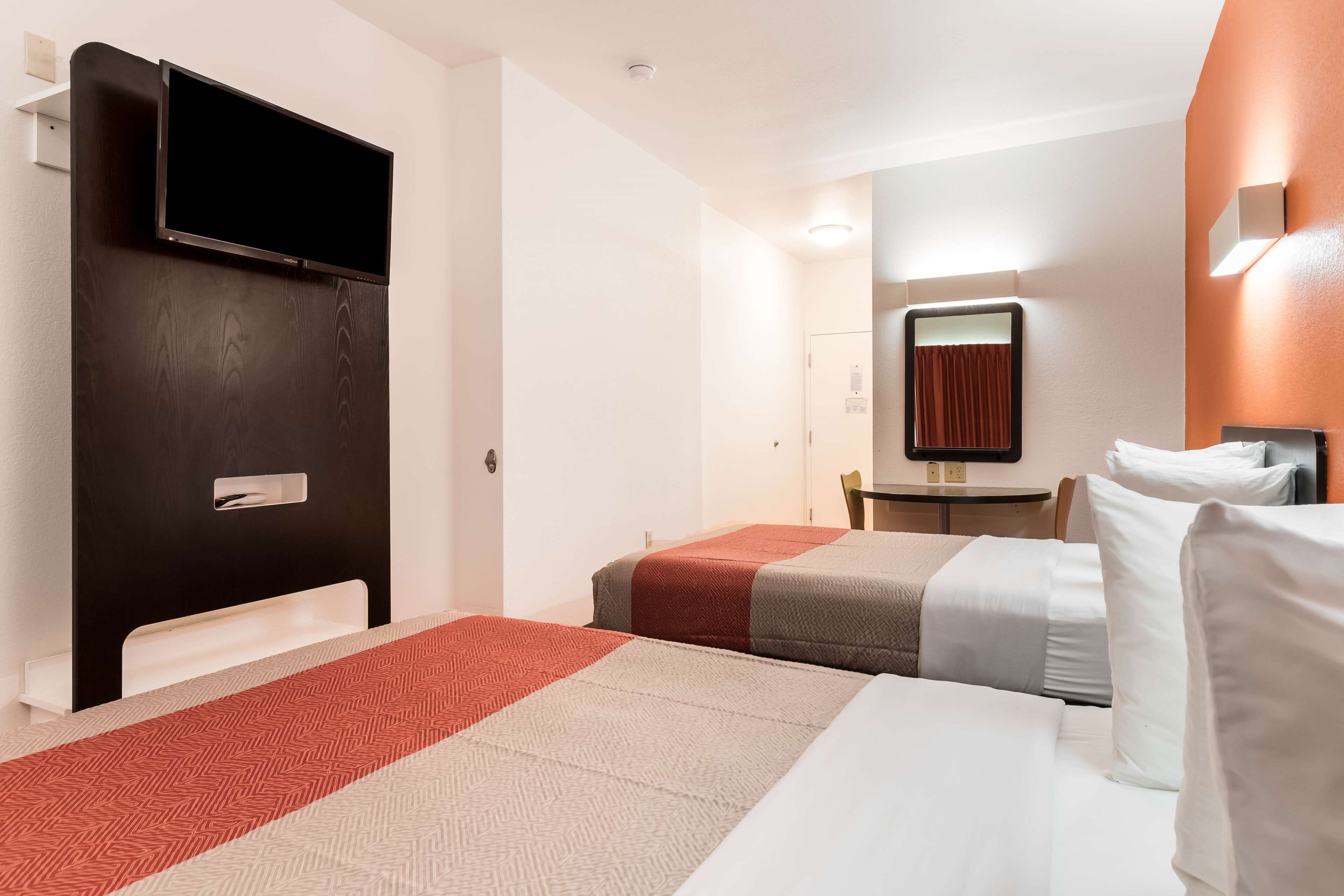 A hotel room with two beds, a TV mounted on a dark wooden panel, a small table with a mirror, and a red curtain.