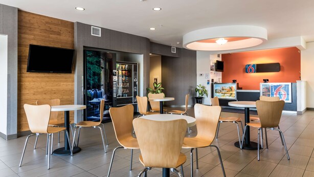 A modern cafe with round tables and wooden chairs, a TV mounted on the wall, and a counter with a sign. - Motel 6 Wilkes Barre, PA - Arena (Wilkes-Barre)