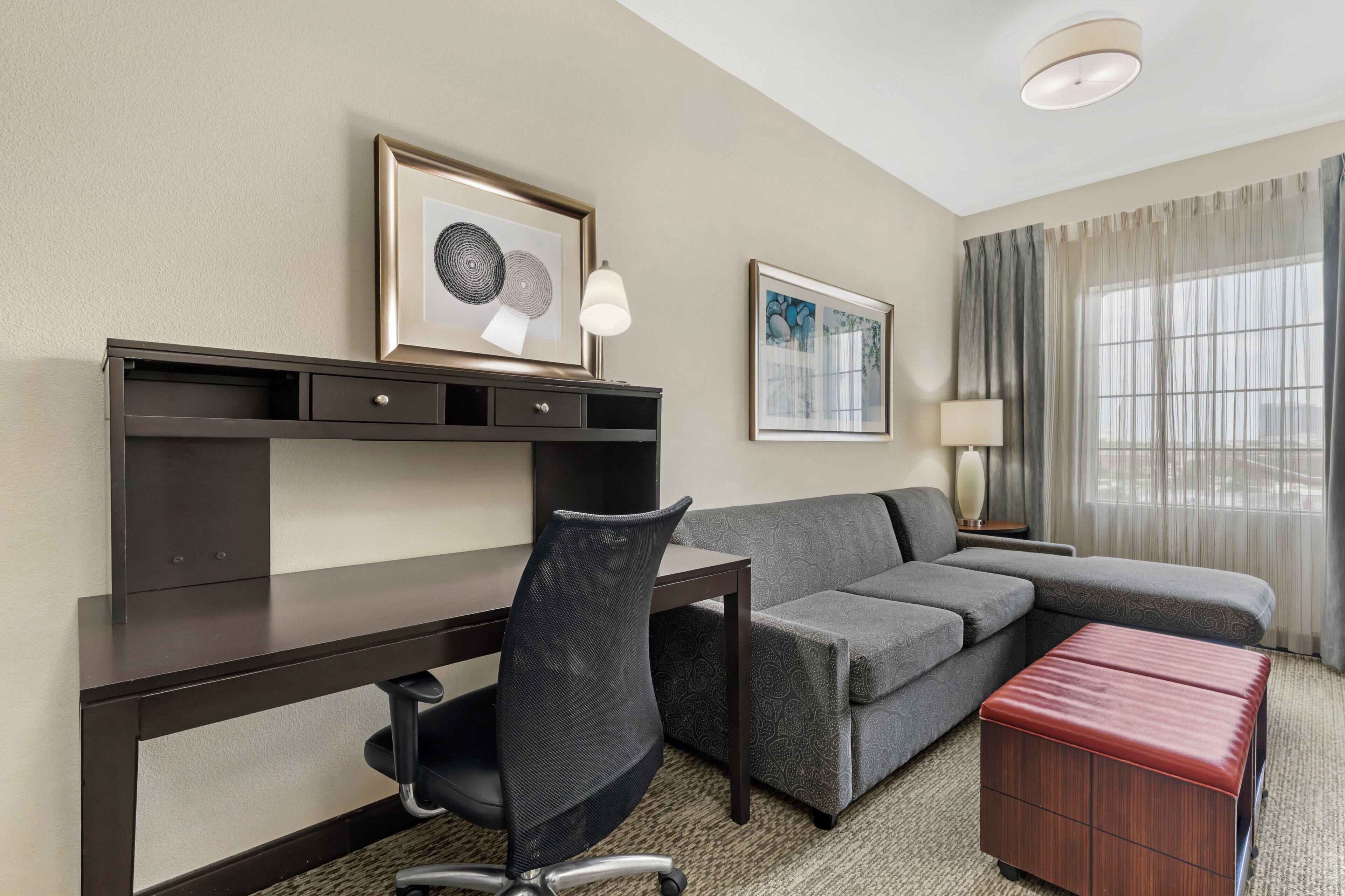 A hotel room with a desk, a cushioned chair, a sofa, a red ottoman, and two framed artworks on the wall.