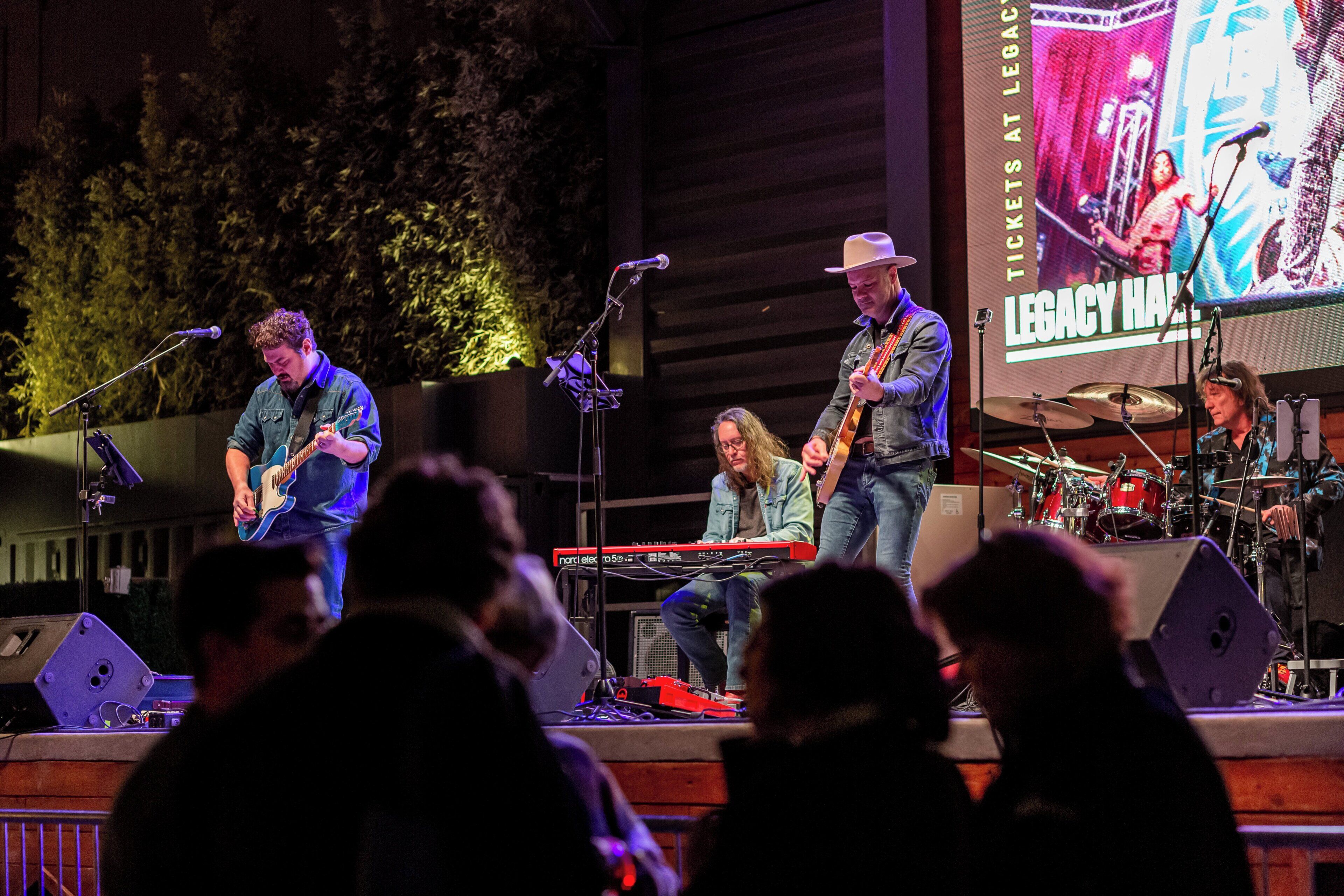Een band treedt op in een theater met een groot scherm waarop 'LEGACY HALL' staat.