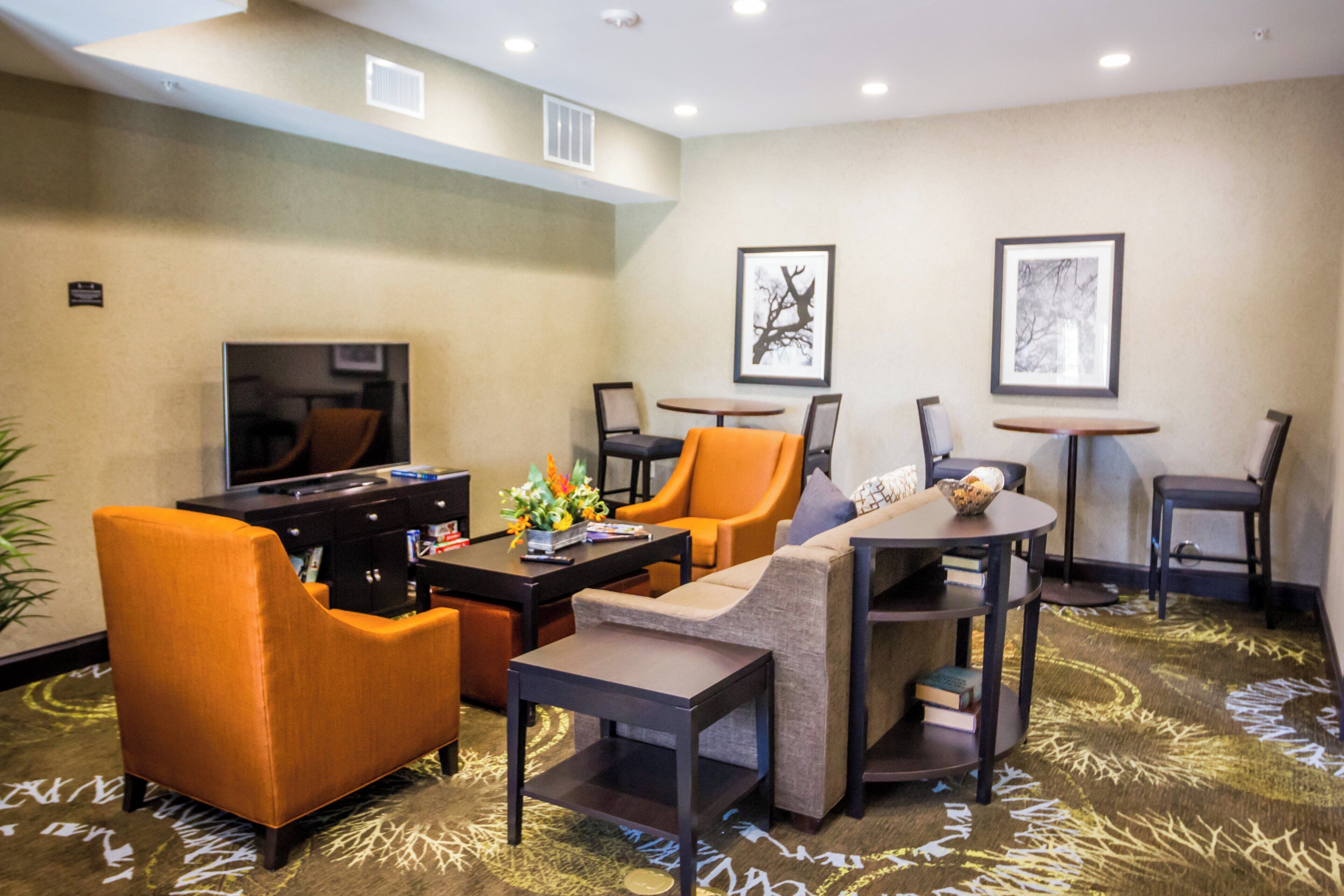 A hotel lobby with a television, orange chairs, a sofa, a coffee table, and framed artwork on the walls.
