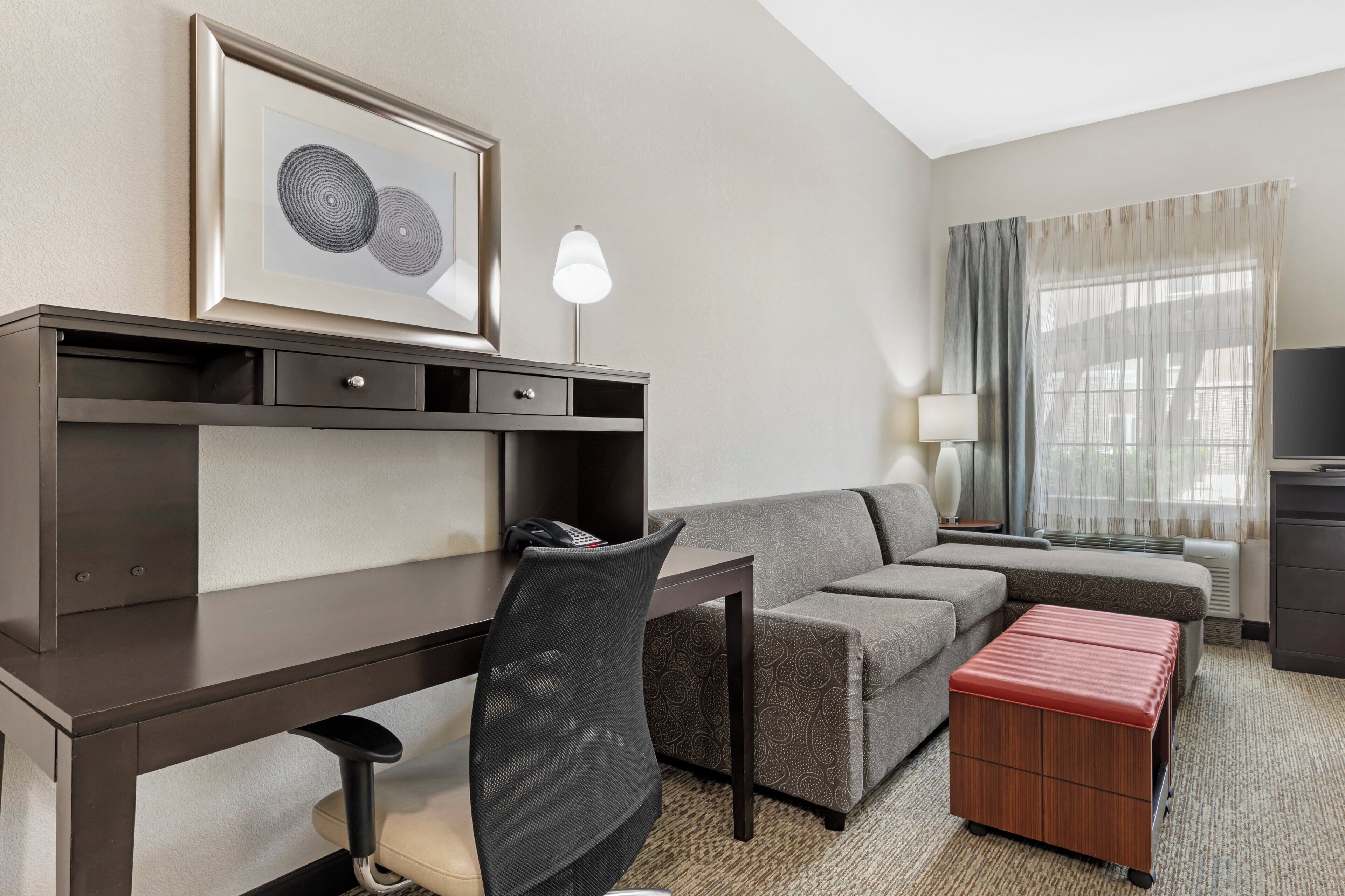 A hotel room with a desk, a chair, a sofa, a small ottoman, a television, and a window with curtains.
