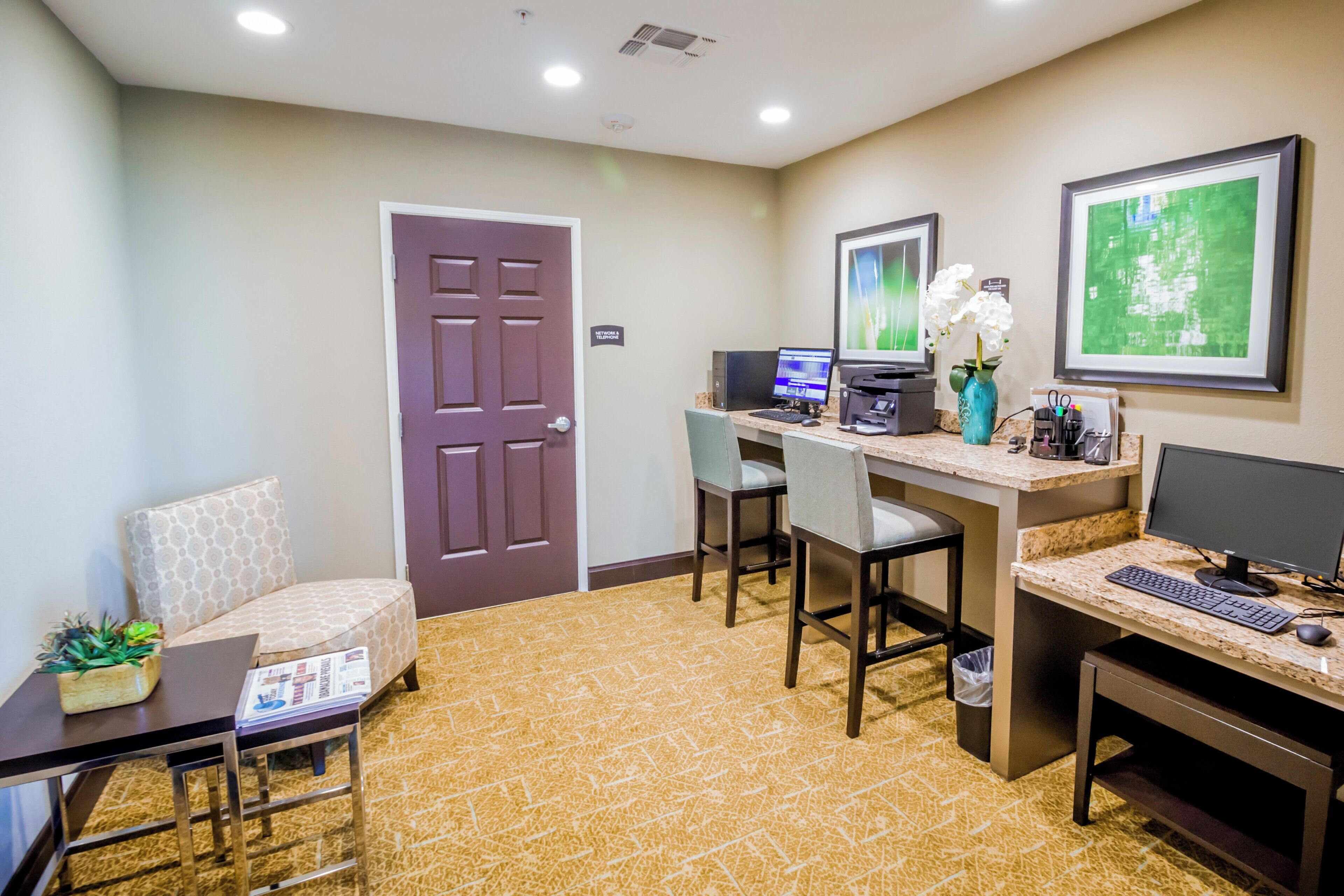 Habitación de hotel con escritorio, computadora, sillas, una pequeña mesa con una planta y una puerta.