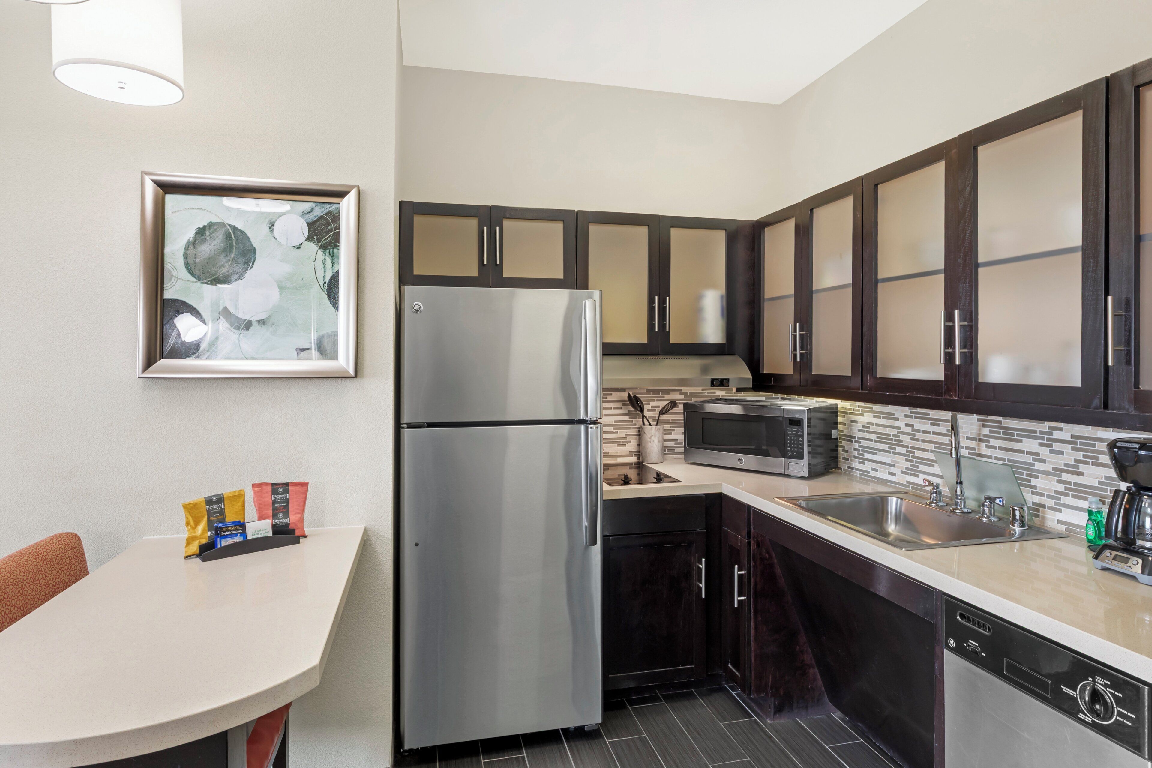 A modern kitchen with a refrigerator, microwave, and dishwasher. There is a dining table with chairs and a framed picture on the wall.