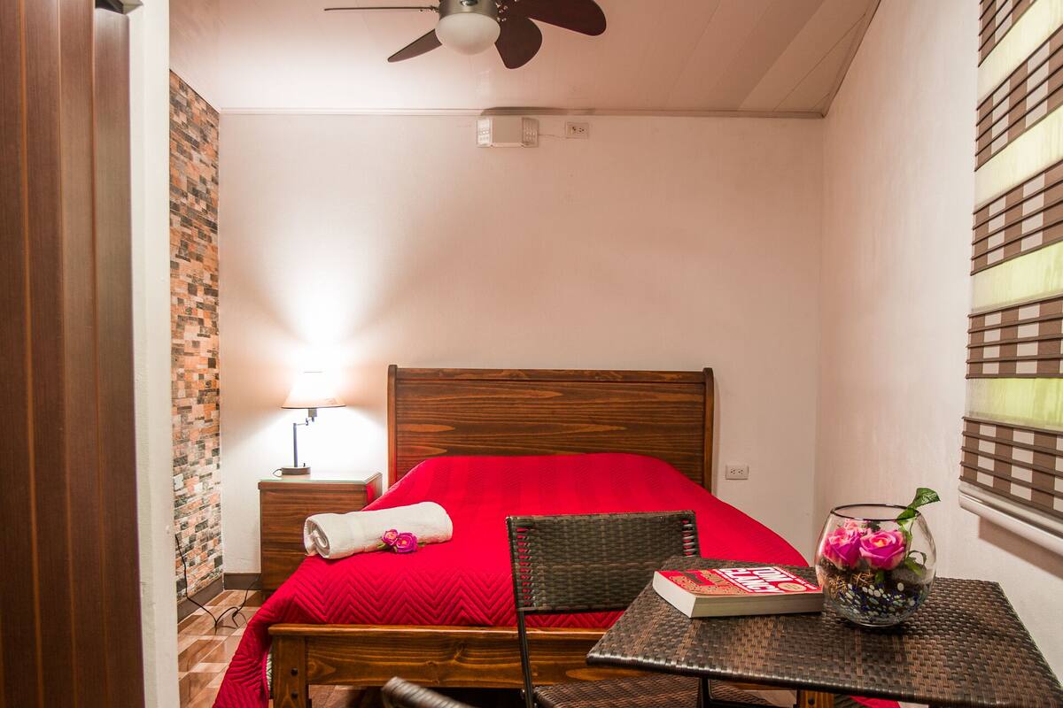A hotel room with a bed, a table with chairs, a ceiling fan, and a window with blinds.