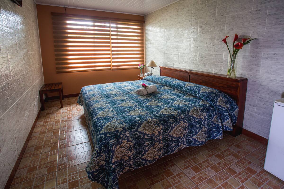 A bedroom with a patterned bedspread, a wooden headboard, a bedside table with a lamp, and a vase with flowers.